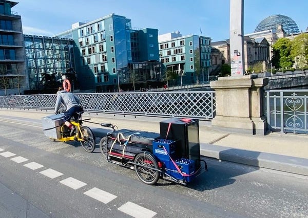 Ein vollbeladenes Cargobike zieht einen mit Kühlschrank, Getränkekisten und Standaufbauten beladenen Anhänger über eine Brücke in Berlin an einem sonnigen Morgen.