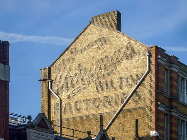 A fading painted sign (ghost sign) on an elevated gable end wall. It is basking in the warm glow of morning sunlight and its fading letters have the word 'Warings' running diagonally across the entire width of the wall in cursive lettering. Under this in block letters are the words 'Wilton Factories'.
