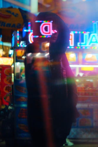 A hooded person is seen through a reflection in front of bright colorful lights from a bright food cart.

The person appears feminine. They have a black hood over their head. Their hands are in their pockets.

The reflection is not perfect; lights from the food cart are overlaid on top of the reflection of the person, creating a mess of lights over their dark silhouette.