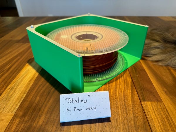 a photograph of a single filament stacker with a spool of copper colored filament in it. The Stacker is green except for the top which is grey. In front of it is a piece of paper that reads "Shallow" for Prusa MK 4. All of this sits on a wooden table.

on the right edge of the photo you can see the fluffy tail of my cat. ;) 