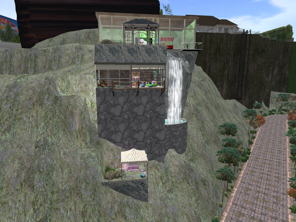 A Second Life house that's basically a large vertical cliff with a waterfall, and there's a building on top of the cliff as well as another integrated into the cliff. There's a little platform at the bottom with a tent and an ice cream counter inside.