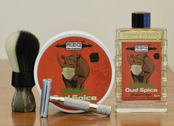 A shaving brush with a camo handle and a synthetic knot, half white and half black, stands next to a but of shaving soap with a reddish-brown label on which is a black line drawing of a rhinoceros wearing a white surgical face mask. the name "Oud Spice" is in cream-colored black letters at the bottom. Next is a rectangular transparent glas bottle with a black and the same label with "Aftershave & Cologen" added in script at the bottom. Lying on its side in front is a slant DE razor with a stainless-steel bulldog handle.