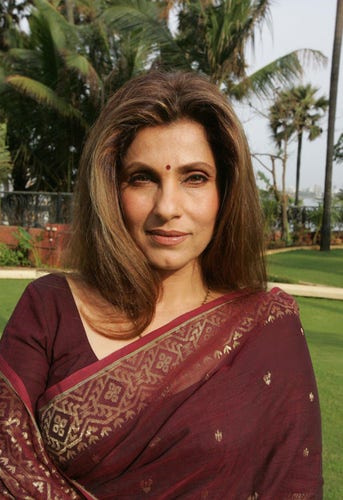 medium close-up shot of zdimole Kapadia. She's standing somewhere outside. There's grass and trees visible behind her. She's wearing a maroon choli blouse and a sari in the same color with a gold border. Her long brown hair is down. She's looking into the camera. she's gorgeous 
