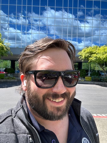 @urda@urda.social smiling In the sun with his sunglasses on. A mirror like finish is on a building behind him, reflecting a beautiful blue sky.