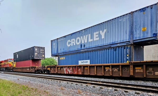 Peeking out of a small wooded area onto the "no trespassing" area adjacent to a train track as a freight train passes, with a colorful orange and yellow engine pulling double stacked box cars in many colors.