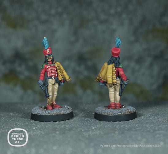 wargame model. Chap with long hair and a handlebar moustache wearing a tall military style hat tunic and Prussian jacket, holding a riding crop. The hat and tunic are in a cerise-crimson and the jacket is in yellow. He has a pair of round glasses on with green lenses.