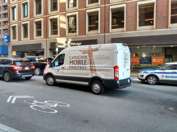 Stuck in Boston street traffic, a white van that says "Capuchin Mobile Ministries" tries to go somewhere. 

There a brown graphic that looks like a robed male cartoon figure with arms outstretched in a crucification look. He's standing kind of on the back wheel well. 