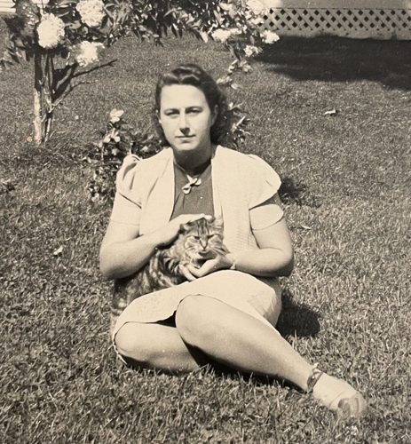 Black and white photo of a white woman sitting on a sloping lawn, looking at the camera and cradling a longhaired tabby cat in her lap.