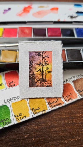 Tiny painting leaning on a small watercolor palette. The painting shows a landscape with bright yellow-orange sky reflected in water and trees