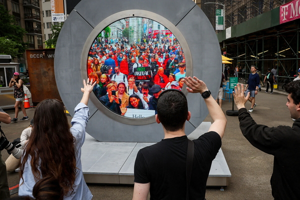 A photo from the linked article showing a group of people in New York City standing in front of "the Portal", a round, gray structure that's a bit taller than an average adult, with thick bezels, and a large screen embedded in the center, showing a crowd gathered in Dublin, waving and taking pictures with their cellphones. One person is holding a sign that says "Free virtual hugs", with each word on a separate line, with each word written using a different color of the Irish flag (green, white, and orange, from top to bottom).