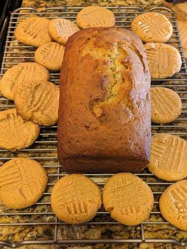 Golden-brown peanut butter cookies surround a loaf of brown banana bread on a cooling rack. Situated on a kitchen counter.