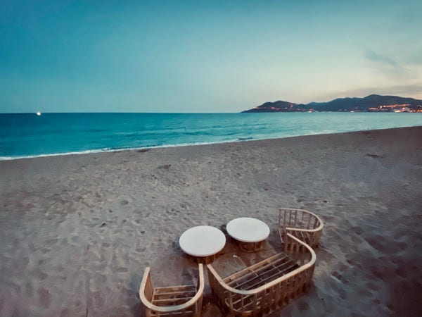 Two wicker chairs and a small table on a sandy beach with the sea and distant hills of the Esterel in the background at dusk. 
