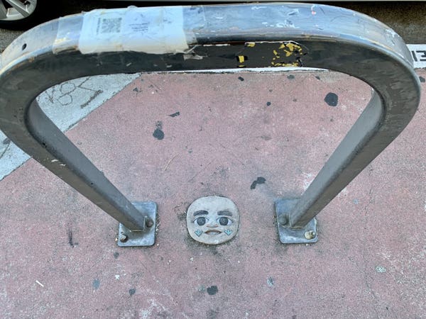 A sculpted clay face glued to the sidewalk beneath a bike rack. There is also a grey bike locked to the rack. The face is about 4 inches wide and appears to be smiling. It has been painted, but the paint mostly faded. It has big loving eyes, and it's mouth is partially open and smiling.