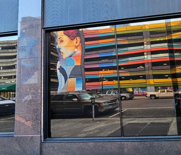 In a clear glass window, directly across the street from a mural, the reversed reflection of a seven story parking garage with corner, "tower-like" stairwells has been brightly painted with a rainbow-like mural also containing portraits of two female faces. The garage levels have bright shades of yellow, orange, blue, purple, red, and white in swirling patterns. The stairwells has a side view and front-facing portrait of an unknown woman's face, in multiple shades of blue, red, white, and orange.