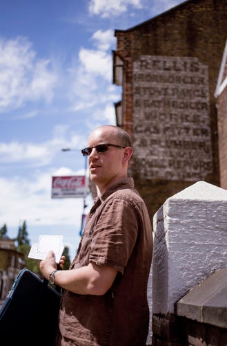 Man in a short-sleeved brown shirt and sunglasses talking in front of a wall that has the fading remains of a painted sign on it.