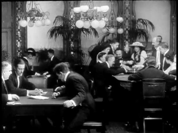 There are two tables with people around them, gambling. Most people in both desks are men, but we can see a woman too. They are indoors.