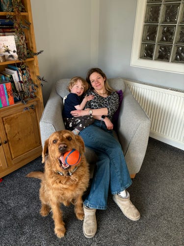 My wife and son together in an armchair, with the dog sitting by her feet with his ball in his mouth, all looking at the camera.