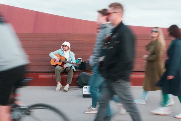 Street performer sitting with an electro-acoustic guitar plugged into a portable speaker. In front of him, blurry silhouettes of moving people can be seen.