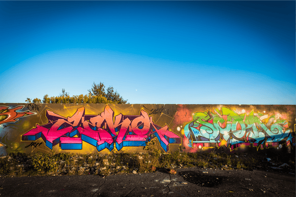 This image showcases a vibrant graffiti wall under a clear blue sky. The artwork is rich with various hues—predominantly shades of pink, blue, green, and yellow—and is full of complex shapes and patterns that make up stylized letters and forms. Sun flare dots the photo, adding a lens flare effect to the lower part of the frame. The graffiti is bordered by a line of green foliage at the top, while the ground in front shows signs of wear and sparse vegetation. The presence of the moon faintly visible in the sky suggests this photo was taken during the day close to dusk or dawn.