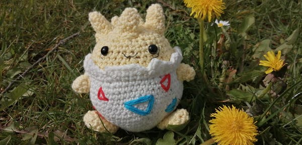 A crocheted Togepi sitting on a meadow next to a blooming dandelion