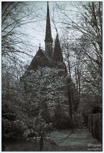 Black and white picture of a roman catholic chapel taken from the graveyard. The chapel has a bell tower with a very steep spire, a cross sitting on the other end of the roof and another, less steep, spire on a lower part of the building. It is quite dark against a clear sky and partially obscured by trees catching the sunlight.