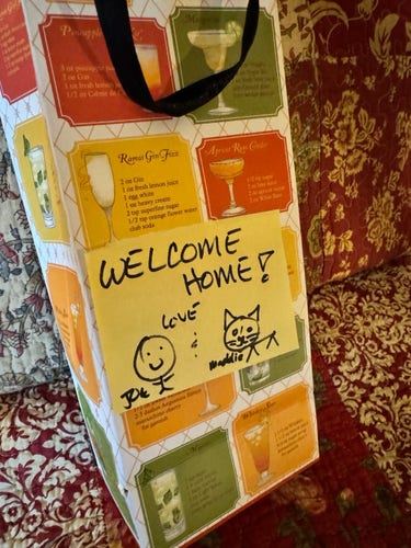 A gift bag with cocktail recipes printed on it and a "WELCOME HOME!" note attached, decorated with handwritten signatures, a smiley face, and a dog drawing.