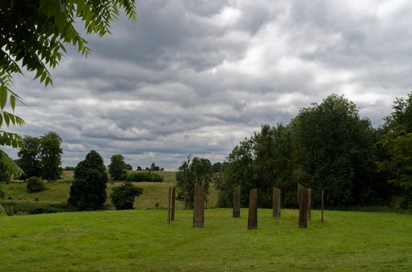 A group of nine standing stones in a field of grass, with trees on the right-hand side. The sky is grey and cloudy.
