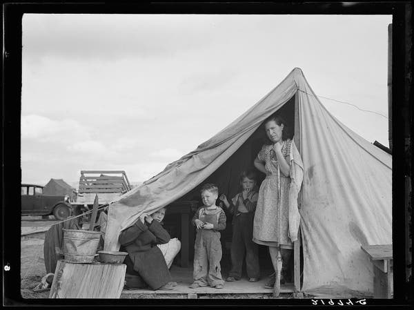  The image depicts a family living in a tent during the Great Depression era. There are five individuals present: an adult woman and four children. The woman is standing on what appears to be the entrance of the tent, while the children are sitting or standing nearby. They are dressed in casual clothing suitable for outdoor activities or work. The setting suggests a rural environment with a backdrop that includes other tents and vehicles. The overall mood of the image conveys a sense of resilience despite the hardships faced by the family during this period.