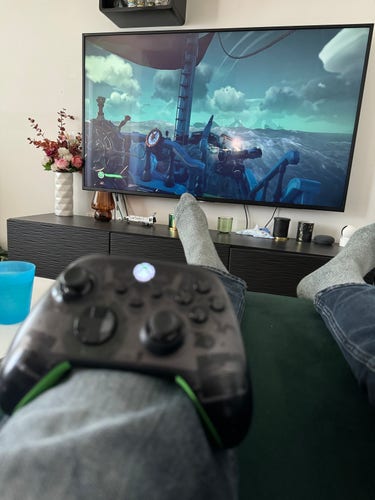 Person playing a video game with a controller, visible legs and feet resting on an ottoman, with the game on the TV screen in the background.