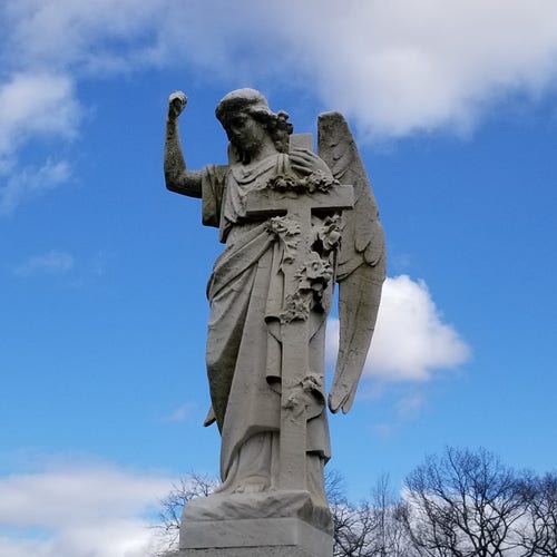 Stone angel in Orchard Street Cemetery in Dover, NJ. Right arm raised toward heaven; left arm holding cross with flowers wrapped around. Probably Gabriel. Blue sky with some puffy clouds and bare tree branches in the background.