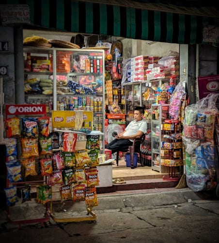 A late evening photo of a shop proprietor kicking back on his phone amid the shelves and racks of packed goods in his Hanoi tạp hóa (local neighborhood convenience store). He is in a white, short-sleeved polo and dark trousers. He sits on a stack of three plastic chairs looking down at his phone. Out front on the pavement, we see two branded snack chips racks ("Orion" & "Lays") to the left of the entrance. On the right is another unbranded rack wrapped in plastic. Inside is like a greatest hits of tạp hóa goods: instant coffee, canned & bottled soft drinks, instant noodles, toilet paper, snack cakes, coca-cola bottles, candy, cigarettes.