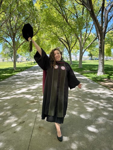 A smiling woman in graduation attire holding up her cap, standing on a tree-lined path.