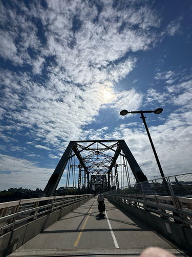 A person biking enters onto the Alexandra bridge in Ottawa Ontario as it is closed to people driving cars. There are white puffy clouds over a very blue sky in the background 