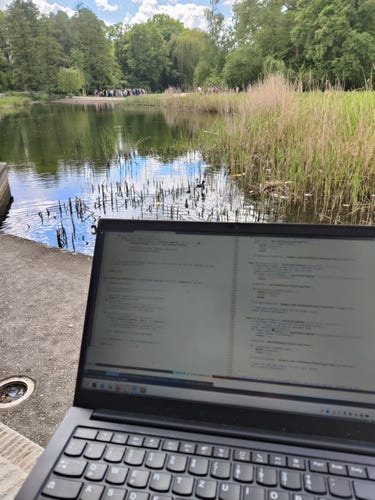 Picture of a park with some water on the background and a laptop on the foreground with some code
