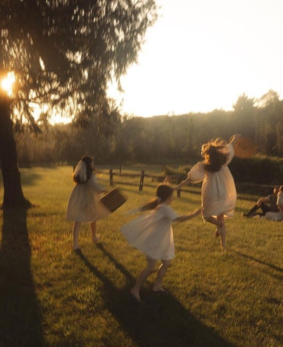 
Photography. A color photo of a group of girls dancing in the sunlight. Three young girls in simple white dresses and white stockings dance across a green, fenced-in meadow. They are seen from behind. Their hair is flying, one girl is in the air and spreads her arms. Another girl is in motion and the third is wearing a white veil and holding a suitcase in her hand as she walks. On the left is a deciduous tree with the sun shining through its branches. On the far left is a man with a small child watching them. The picture has a warm atmosphere and gives the impression of coming from a bygone era. It is part of a series of photographs taken at a wedding.