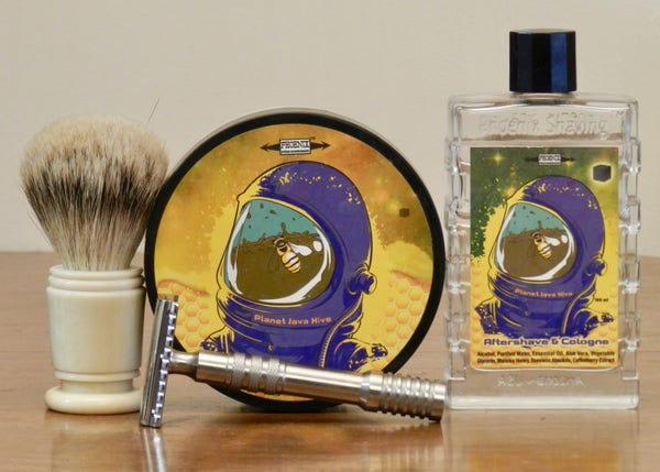 A silvertip badger shaving brush with cylindrical ivory-colored handle stands next to a pub of shaving soap that shows the purple helment of a spacesuit with a yellow background. The helmet is labeled "Planet Java Hive" in gold and the faceplate reflects or reveals a honeybee floating on coffee. Next is a rectangular clear-glass bottle with the same label. Lying on its side in front is a stainless steel razor.