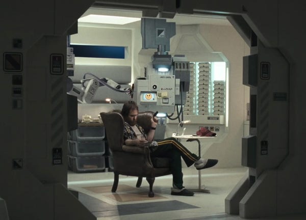 Sam Rockwell as Sam Bell in the movie “Moon” (2009), sitting in a leather chair with his legs casually crossed, in a sterile white room of a moon base. He’s flanked by stacks of packaged meals and a ceiling mounted AI robot with a emoji face, frowning slightly. 