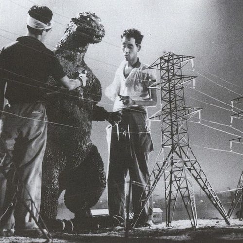 A photo of Godzilla with two men on both sides, getting him ready for a scene in the movie. They are standing in a movie set that shows miniature buildings and power lines.