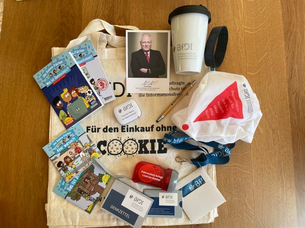 Merchandise des BfDI
Automatisch generiert:
A variety of promotional materials laid out on a wooden surface, including informational comic books, pamphlets, a mug, a fabric tote bag, pens, a lanyard, a face mask, and small gadgets with branding related to BfDI.