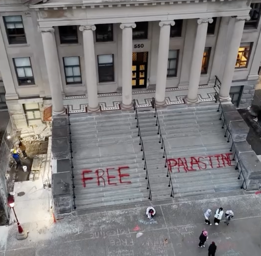 Screen shot from music video showing drone view of a university area, where student’s have written“Free Palastine” [sic] on the stairs in blood red paint.