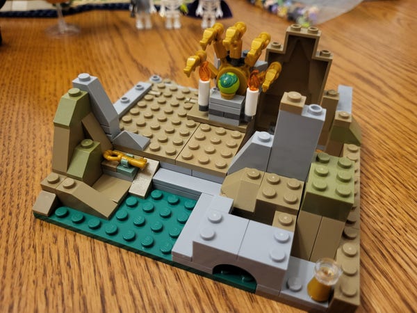 Bag 19 of the Lego Dungeons & Dragons set, completed. The main feature of this portion is a shrine of some kind with a green sphere, two candles, and five golden arms holding small dragon heads. The shrine rests on a small platform surrounded by brown, gray, and moss green pieces that may represent rocks and dirt.