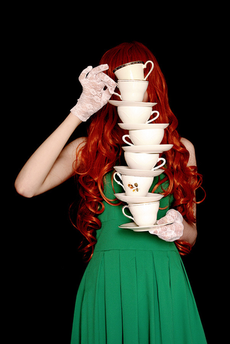 A woman with long red hair in a green dress is holding a staple, one above the other, of coffee cups, so high, it is covering her face.

©Vicky Martin