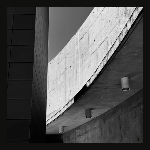 This image presents a modern architectural detail in black and white. It features a stark contrast between a smooth dark panel on the left and a textured concrete wall on the right. The right wall extends over the left, creating an overhang that is punctuated by cylindrical lights recessed into the concrete. These lights are evenly spaced, projecting out slightly from the surface. The shadows cast by the overhang and the geometric precision of the construction emphasize the clean lines and the minimalist aesthetic typical of contemporary architecture. The overall composition plays with depth and perspective, highlighting the angular intersection of these two architectural elements.