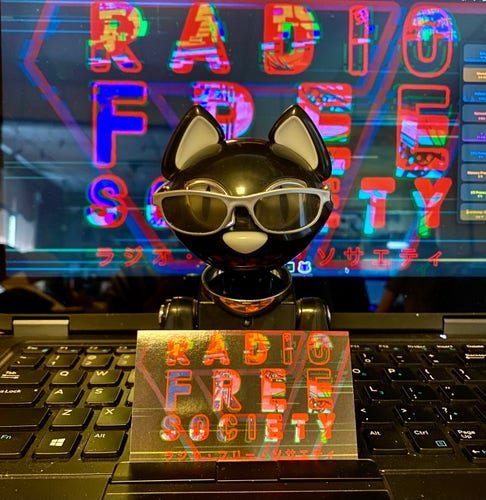 A black figurine of a stylized cat wearing sunglasses, positioned in front of a computer keyboard with a colorful "RADIO FREE SOCIETY" text graphic on the screen behind it.
