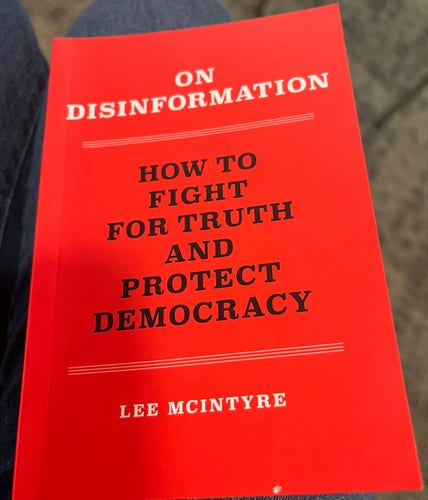 A red book titled "ON DISINFORMATION" with the subtitle "HOW TO FIGHT FOR TRUTH AND PROTECT DEMOCRACY" by Lee McIntyre.