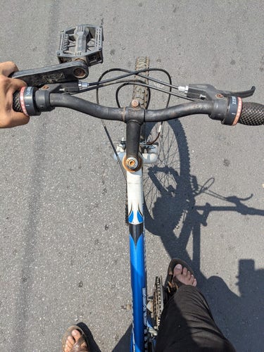 una bicicleta en movimiento vista desde el ciclista a la que le falta el pedal izquierdo, que es sostenido en la mano izquierda junto con el manubrio 
