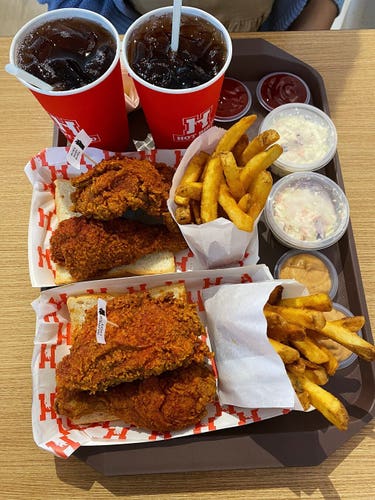 A large plastic tray of foods served on it including 2 paper plates each with a piece of white bread, 2 red hot Fried Chicken pieces placed on top of them, and a paper bag of Cajun Fries. Next to them are several plates of chilli sauce and hot sauce of some kind in yellowish mustard colour, 2 small plastic containers of Coleslaw, and 2 paper cups serving cold Iced Lemon Tea.