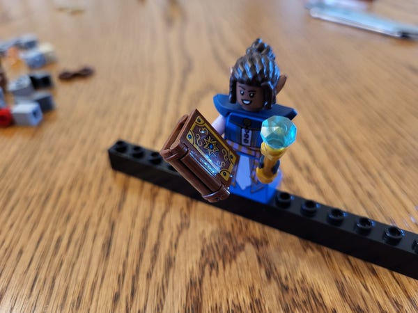 The female elf wizard minifigure from the Lego Dungeons & Dragons set. She has dark skin and hair, and wears blue and purple robes represented by the "dress" minifig piece instead of the usual legs. She also has a piece representing upturned shoulder pads. She carries a gold wand with a blue gem on the end, and a brown spellbook with an elaborate cover. Her expression is wryly amused.
