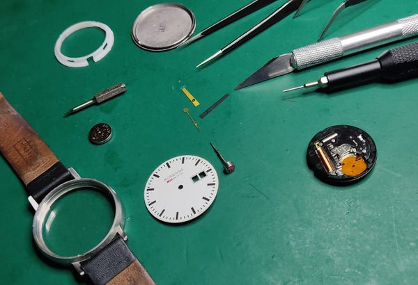 Parts of a quartz wristwatch spread out: case with straps, faceplate, module, battery, hands. Also shows tools: tweezers, cutter, screwdriver 