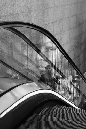 A person riding an escalator up is seen through the glass railing of the escalator, overlaid with a reflection of a black feminine face next to many smaller faces.

The person riding the escalator has short hair. They are mostly obscured by the reflection.

In the background, a stone tile wall extends from the top of the photo down behind the escalator.

In the foreground, the top of the steps from an escalator besides the one the person is riding.